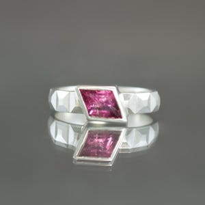 May include: A silver ring with a faceted band and a rectangular, pink gemstone set in the center. The ring is displayed on a reflective surface, creating a mirrored effect. The gemstone is a vibrant shade of pink.