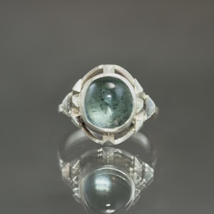 May include: A silver ring featuring a large, oval-shaped, bluish-green gemstone. The ring has a geometric design around the stone and on the shoulders. The ring is displayed on a reflective surface, creating a mirrored effect.