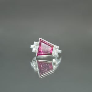 May include: A silver ring featuring a geometric design with a large, angular, pink gemstone. The ring's setting has a modern, architectural style, with small, square accents on either side of the gemstone. The ring is displayed on a reflective surface.