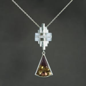 May include: A silver pendant necklace with an Art Deco design. The pendant has a geometric shape with a triangular, multi-coloured gemstone. The necklace chain is delicate and silver, suitable for everyday wear.