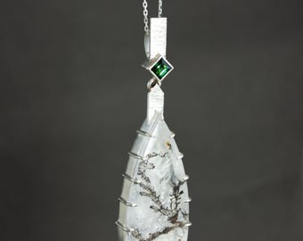 Puede incluir: Un collar con un colgante de plata, que consiste en un cristal de cuarzo transparente con inclusiones negras y un acento de piedra preciosa verde. El colgante tiene forma de triángulo y está suspendido de una cadena de plata.