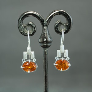May include: Silver drop earrings with an oval, orange gemstone. The earrings have a geometric design above the gemstone and a simple hook closure. The earrings are displayed on a dark metal stand against a gray background.