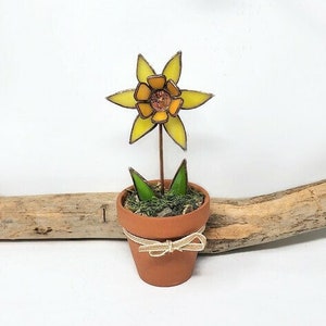 May include: A small, yellow stained glass flower in a terracotta pot. The flower has a six-pointed star shape with a yellow center and orange accents. The pot is decorated with a brown ribbon.