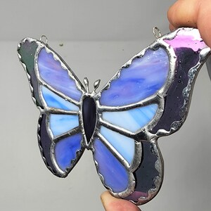 May include: A stained glass butterfly with purple, blue, and white glass pieces. The butterfly has a black center and silver metal framing.