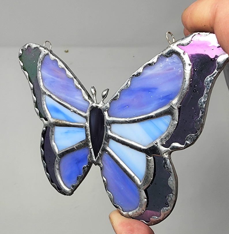 May include: A stained glass butterfly with purple, blue, and white glass pieces. The butterfly has a black center and silver metal framing.