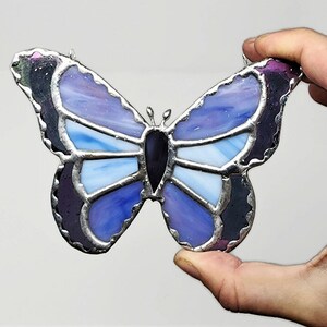 May include: A stained glass butterfly with purple, blue, and white glass pieces. The butterfly has a black center and silver foil accents.