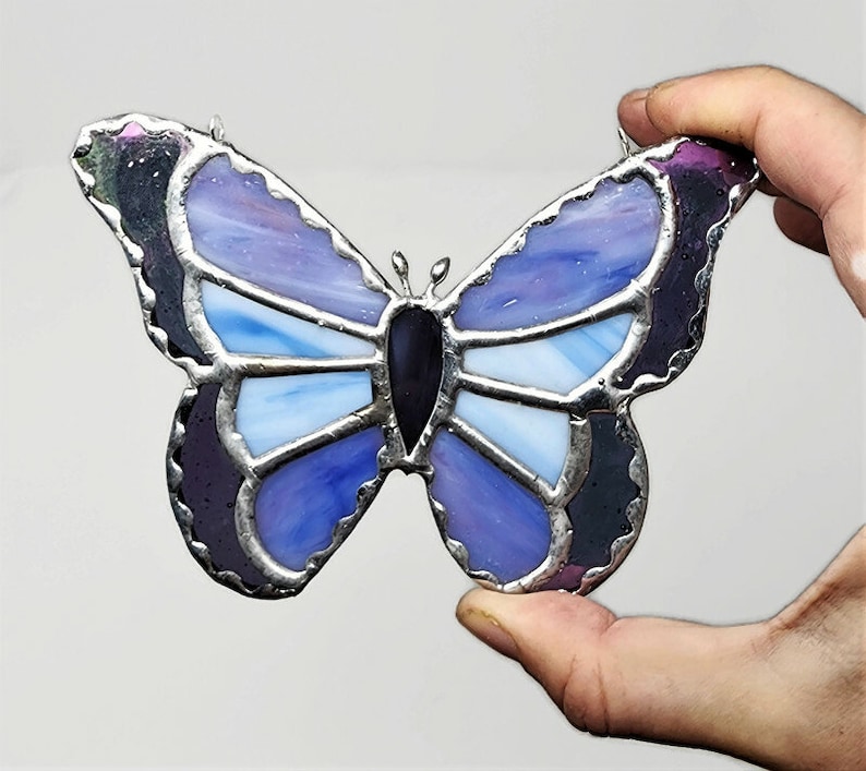 May include: A stained glass butterfly with purple, blue, and white glass pieces. The butterfly has a black center and silver foil accents.