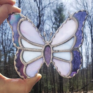 May include: A stained glass butterfly with purple, white, and iridescent glass. The butterfly has a silver frame and is hanging from a string.