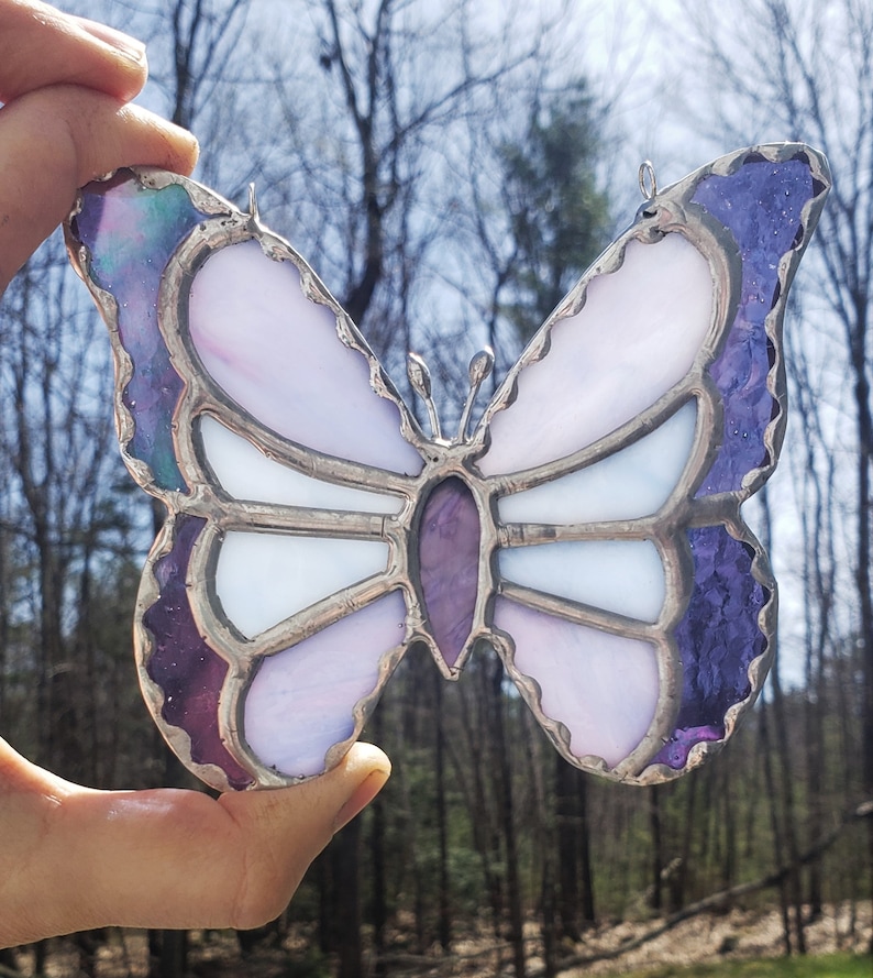 May include: A stained glass butterfly with purple, white, and iridescent glass. The butterfly has a silver frame and is hanging from a string.