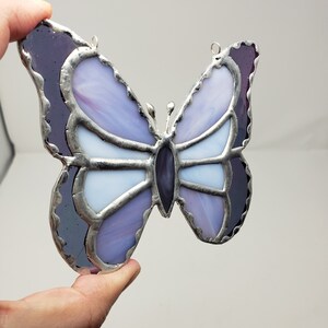 May include: A stained glass butterfly with purple and white glass pieces. The butterfly has a silver metal frame and is hanging from a silver metal loop.