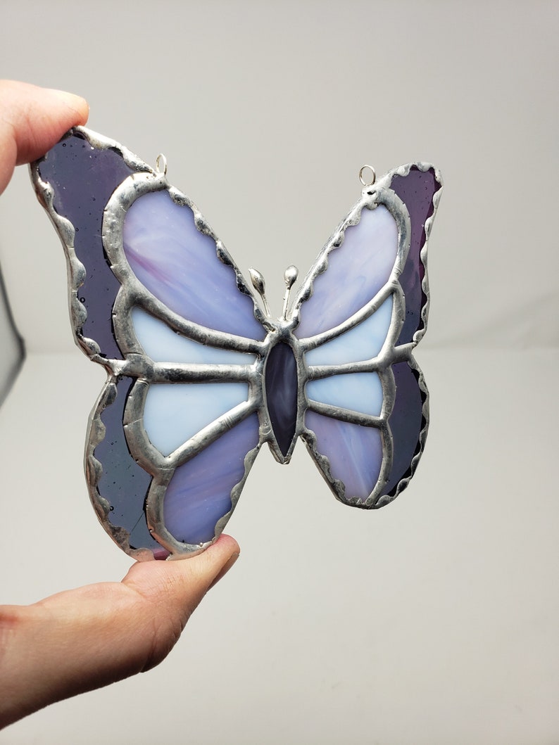 May include: A stained glass butterfly with purple and white glass pieces. The butterfly has a silver metal frame and is hanging from a silver metal loop.
