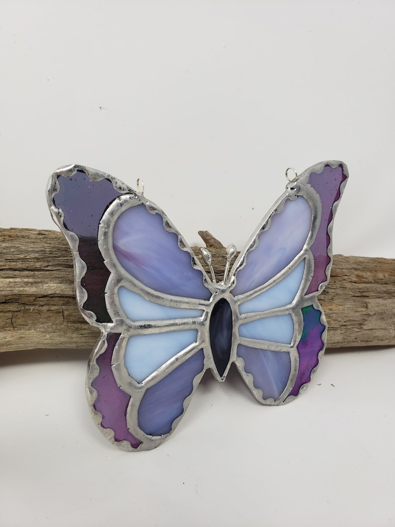 May include: A stained glass butterfly with purple, blue, and white glass pieces. The butterfly has a silver frame and is hanging from a silver wire.
