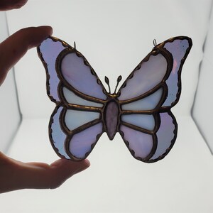 May include: A stained glass butterfly with purple and white glass pieces. The butterfly has a copper wire frame and is a decorative hanging ornament.