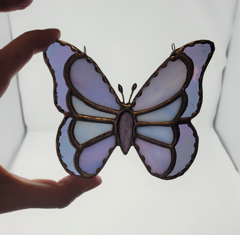 May include: A stained glass butterfly with purple and white glass pieces. The butterfly has a copper wire frame and is a decorative hanging ornament.