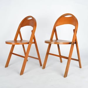 May include: Two wooden folding chairs with rounded backs and seats. The chairs are made of light brown wood and have a simple, classic design.