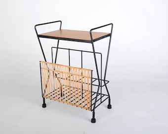 Puede incluir: Un revistero de metal negro con un estante de madera y una cesta de mimbre tejida. El revistero tiene un diseño geométrico y se apoya en cuatro patas negras.