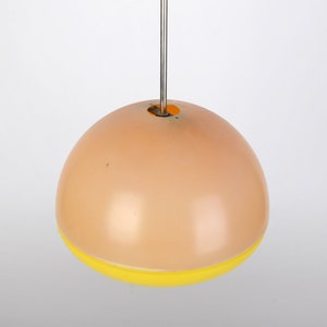 Może przedstawiać: Okrągła, brzoskwiniowa lampa wisząca z ż&oacute;łtym pasem wok&oacute;ł dolnej części. Lampa jest zawieszona na cienkim metalowym sznurku.
