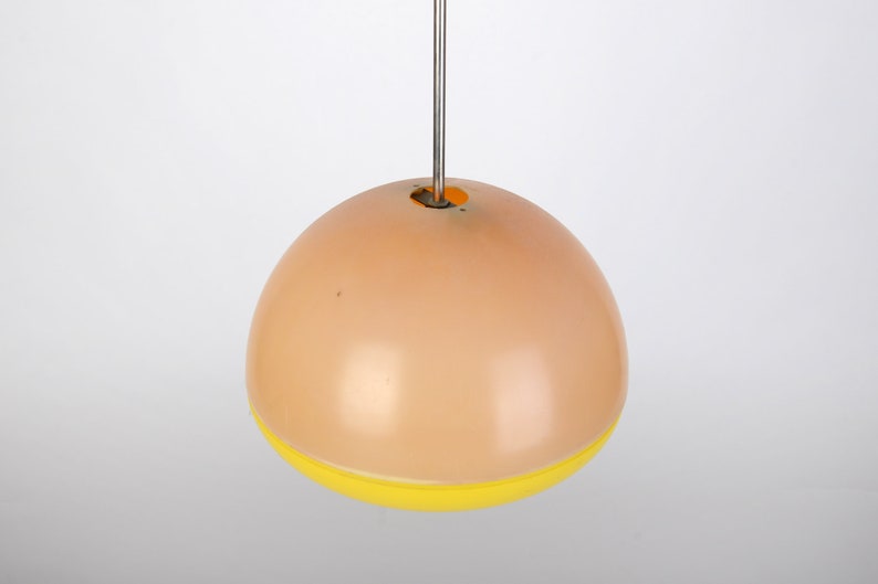 Może przedstawiać: Okrągła, brzoskwiniowa lampa wisząca z ż&oacute;łtym pasem wok&oacute;ł dolnej części. Lampa jest zawieszona na cienkim metalowym sznurku.
