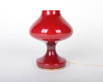 May include: A red glass table lamp with a wide, rounded base and a flared shade. The lamp has a gold-coloured base and a white cord.