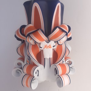 K&ouml;nnte beinhalten: Eine handgefertigte Kerze mit einem gestreiften Design in Blau, Orange und Wei&szlig;. Die Kerze ist in Form eines Baseballtrikots mit dem Wort "Mets" auf der Vorderseite geformt.