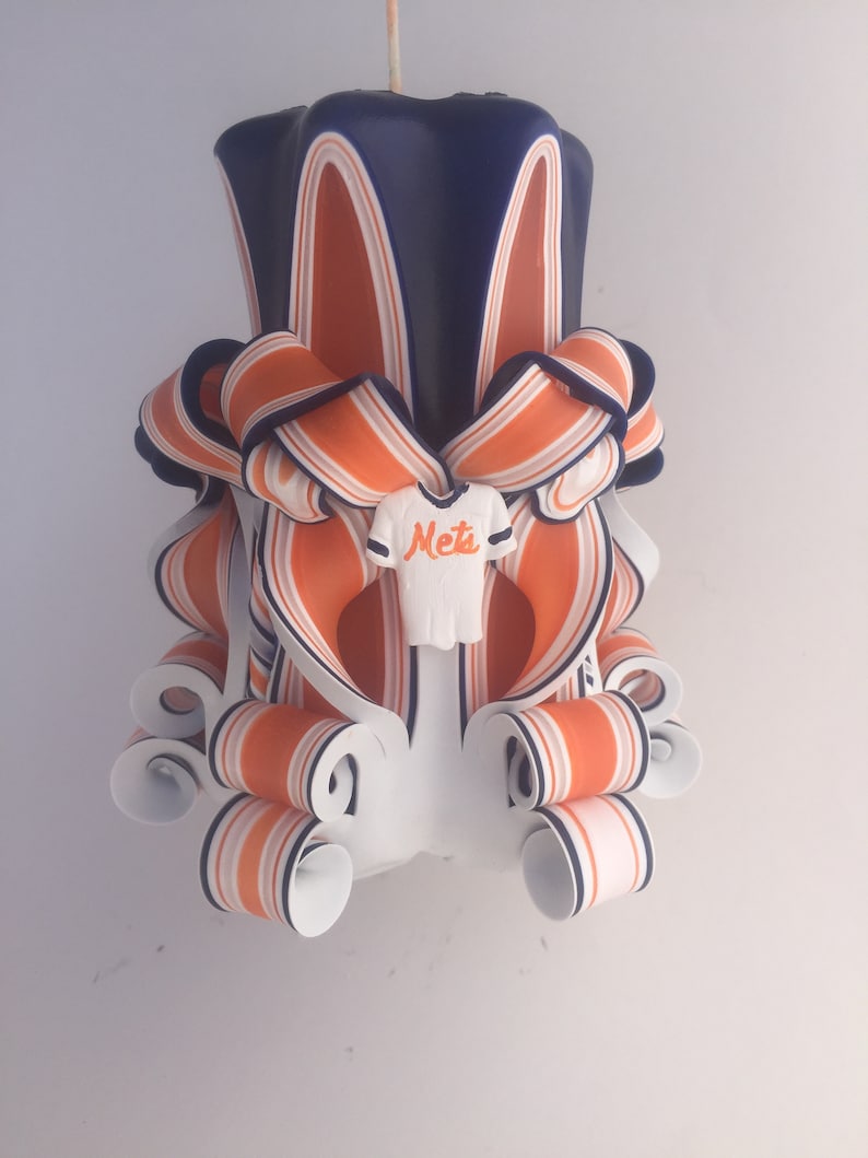 K&ouml;nnte beinhalten: Eine handgefertigte Kerze mit einem gestreiften Design in Blau, Orange und Wei&szlig;. Die Kerze ist in Form eines Baseballtrikots mit dem Wort "Mets" auf der Vorderseite geformt.