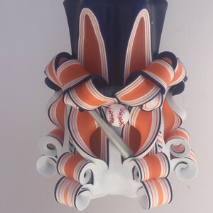 K&ouml;nnte beinhalten: Eine wei&szlig;e, orange und blaue gestreifte Kerze mit einem Baseball- und Schl&auml;ger-Design. Die Kerze ist in Form einer traditionellen S&auml;ulenkerze mit einem dekorativen, gedrehten Design.