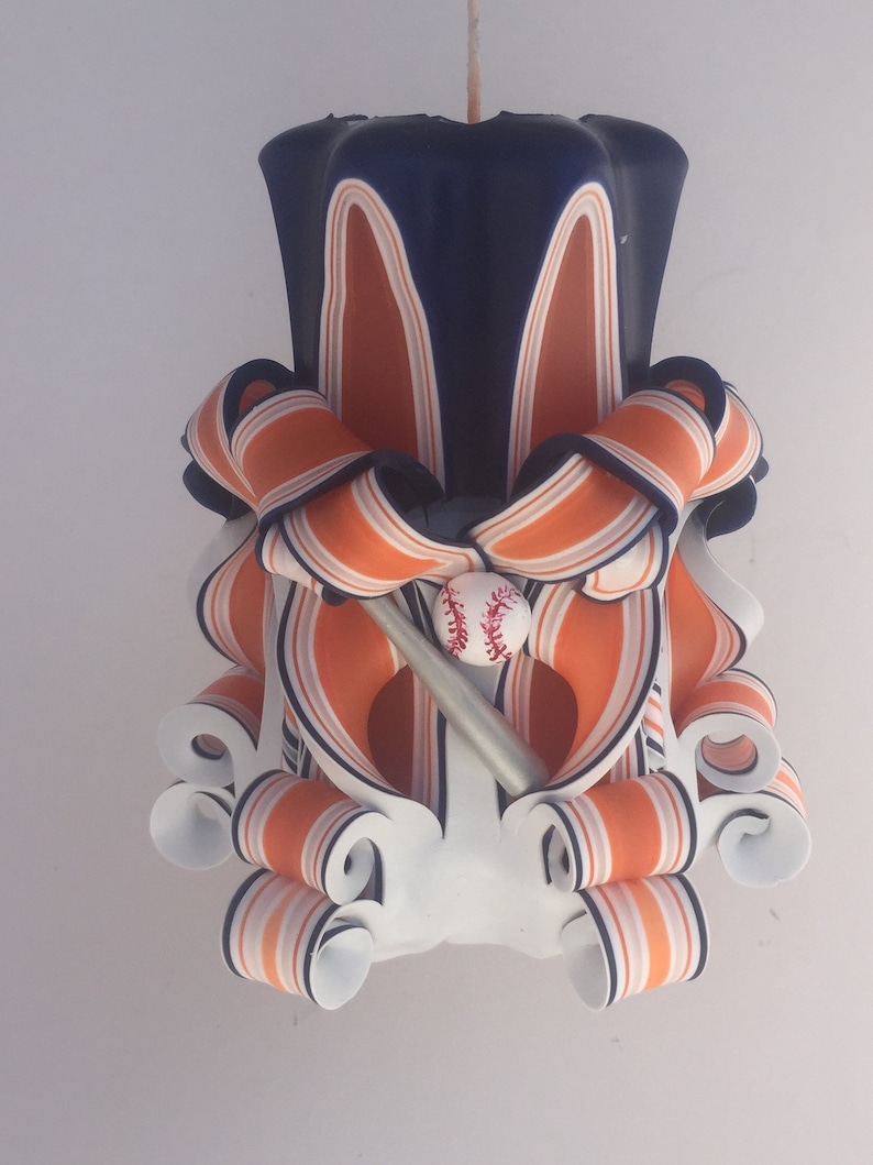 K&ouml;nnte beinhalten: Eine wei&szlig;e, orange und blaue gestreifte Kerze mit einem Baseball- und Schl&auml;ger-Design. Die Kerze ist in Form einer traditionellen S&auml;ulenkerze mit einem dekorativen, gedrehten Design.