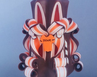 May include: A brown, orange, and white hand-crafted candle with a football jersey design. The jersey has the word "BROWNS" printed on it.