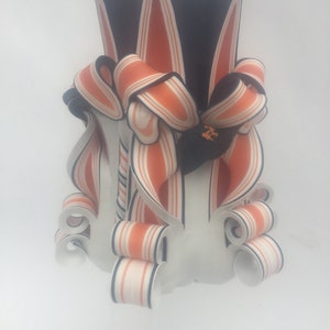 K&ouml;nnte beinhalten: Eine schwarz-orange gestreifte Kerze mit wei&szlig;em Sockel und kunstvollen Verzierungen. Die Kerze ist mit einem schwarz-orangen Band verziert.