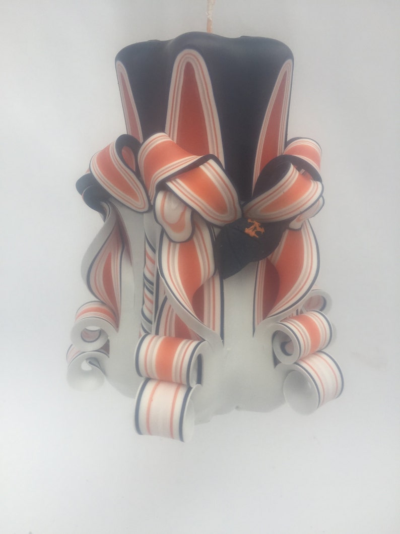 K&ouml;nnte beinhalten: Eine schwarz-orange gestreifte Kerze mit wei&szlig;em Sockel und kunstvollen Verzierungen. Die Kerze ist mit einem schwarz-orangen Band verziert.