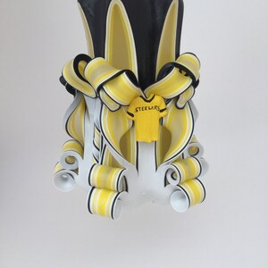 May include: A black and yellow striped candle with a white and yellow football jersey with the word "Steelers" on it. The candle is decorated with yellow and black ribbons.