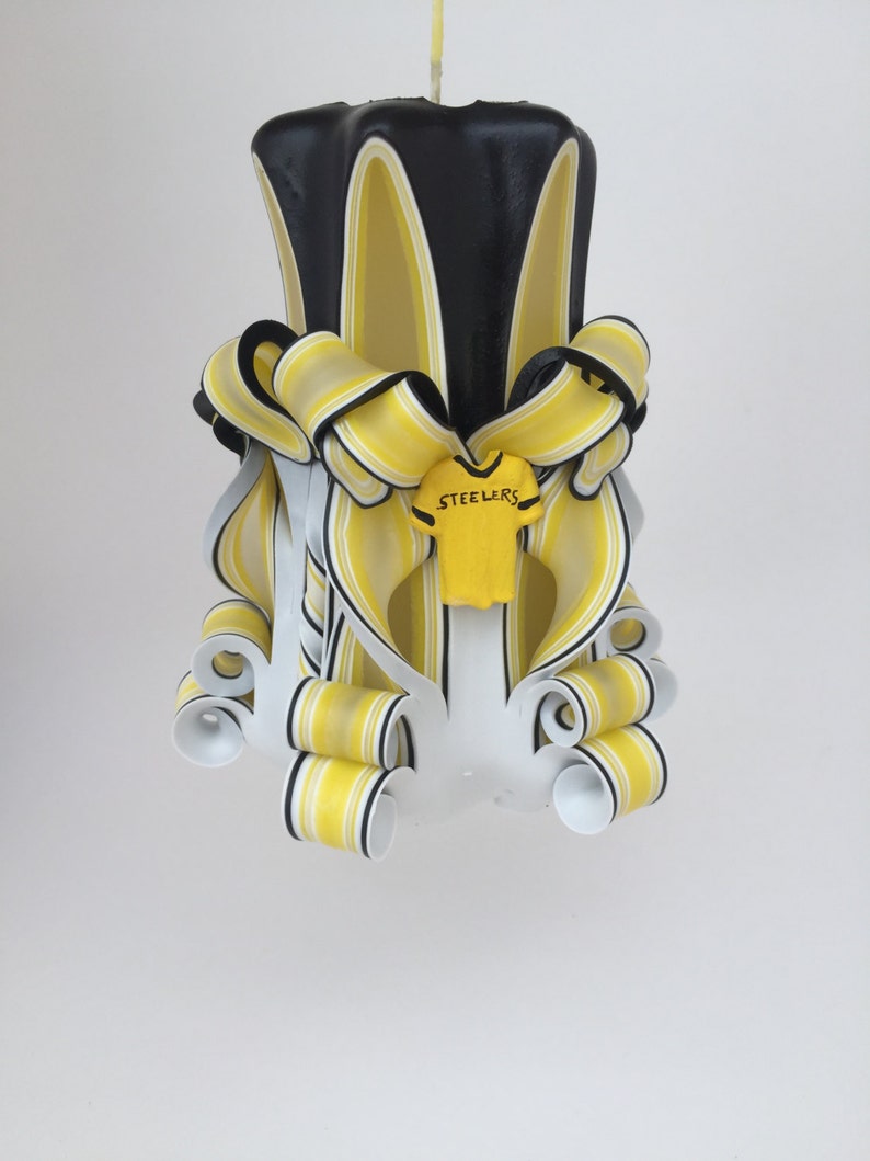 May include: A black and yellow striped candle with a white and yellow football jersey with the word "Steelers" on it. The candle is decorated with yellow and black ribbons.
