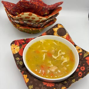 Puede incluir: Un cuenco blanco lleno de sopa, sobre un salvabols de patrón floral. Se ven salvabols apilados con patrones otoñales. La sopa contiene fideos y zanahorias. El salvabols tiene un fondo marrón con diseños florales naranjas y amarillos.