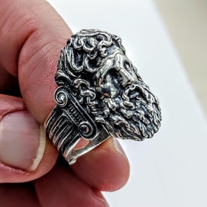 A silver ring depicting the Greek God Zeus, held in the fingers of a hand