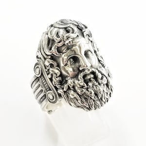 Pode incluir: Um anel de prata com um design detalhado de um rosto de homem com barba e cabelo encaracolado. O anel tem uma banda com um design decorativo.