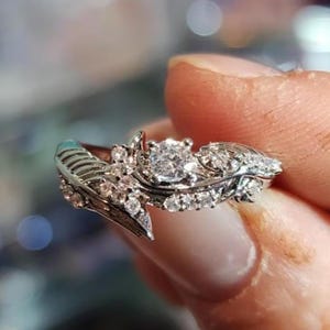 May include: A silver ring with a unique feather design, adorned with multiple small diamonds and a larger, central diamond. The ring's intricate details and sparkling stones create a luxurious appearance, perfect for special occasions.