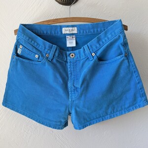 Puede incluir: Pantalones cortos de mezclilla azul con un logotipo de Guess en el bolsillo trasero.