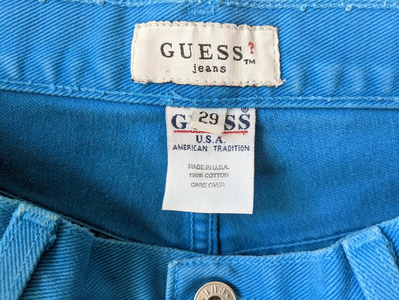 Puede incluir: Una etiqueta de mezclilla azul Guess con el texto "GUESS? jeans TM" y "G29 SS U.S.A. AMERICAN TRADITION MADE IN U.S.A. 100% COTTON CARE OVER"