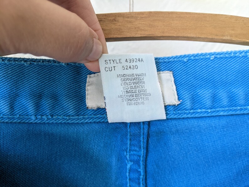 Puede incluir: Una etiqueta de cuidado blanca unida a un par de pantalones vaqueros azules. La etiqueta tiene el texto "STYLE 43924A CUT 52430" e instrucciones para lavar a m&aacute;quina por separado en agua fr&iacute;a, sin lej&iacute;a y secar en secadora a baja temperatura.