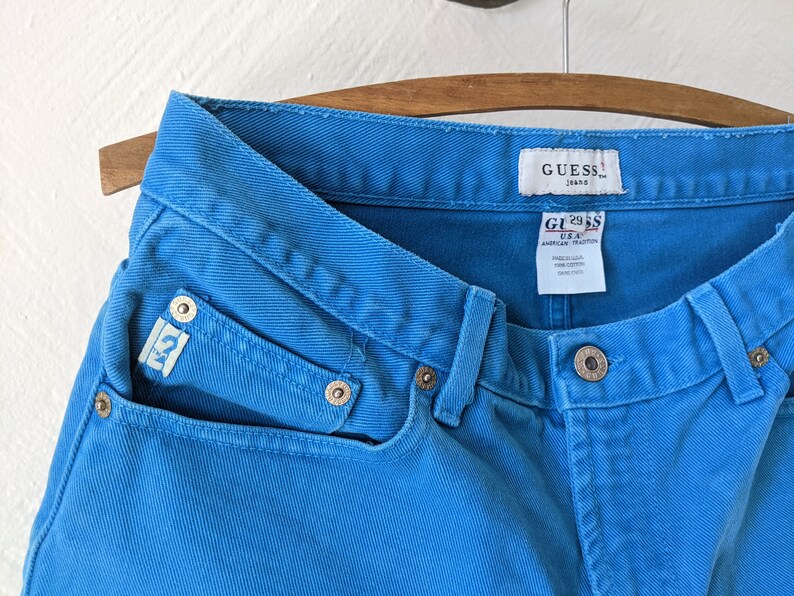 Puede incluir: Un par de pantalones vaqueros azules de mezclilla con una etiqueta de Guess. Los pantalones tienen un lavado azul claro y est&aacute;n hechos de 100% algod&oacute;n.