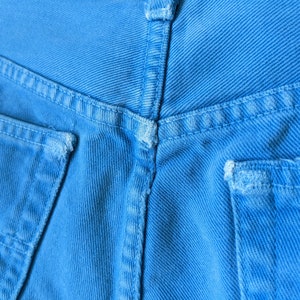 Puede incluir: Primer plano de la parte posterior de un par de pantalones vaqueros de mezclilla azul. Los vaqueros tienen dos bolsillos traseros con costuras alrededor de los bordes. La tela es de color azul claro.