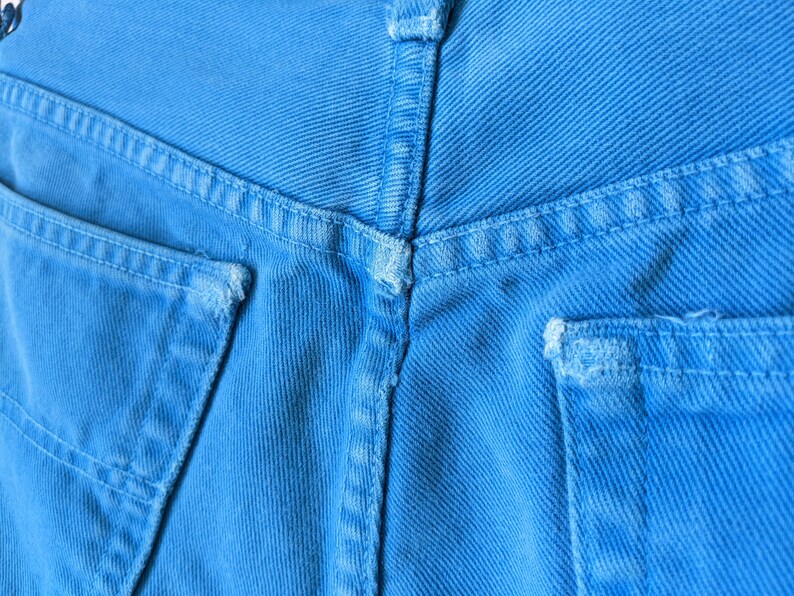 Puede incluir: Primer plano de la parte posterior de un par de pantalones vaqueros de mezclilla azul. Los vaqueros tienen dos bolsillos traseros con costuras alrededor de los bordes. La tela es de color azul claro.