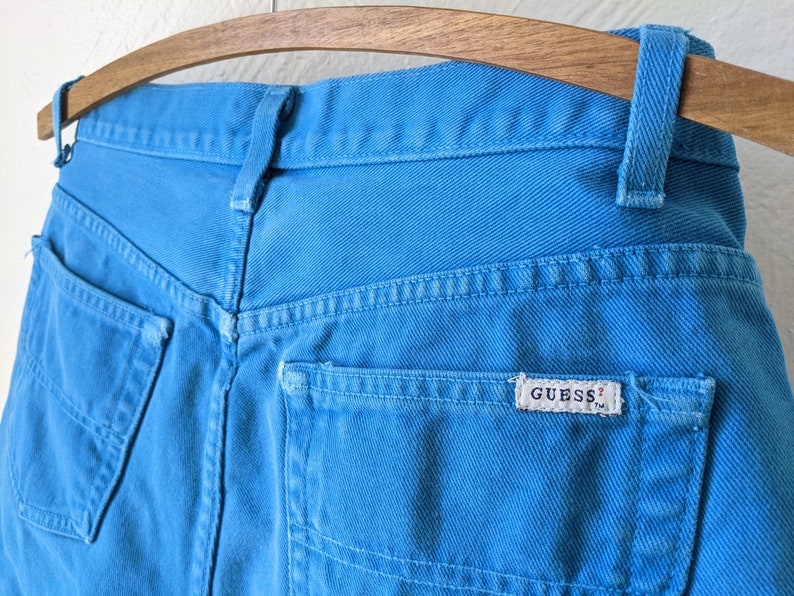 Puede incluir: Un par de pantalones vaqueros azules con un bolsillo trasero que tiene una etiqueta blanca "GUESS?"