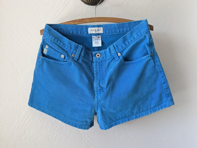 Puede incluir: Pantalones cortos de mezclilla azul con un logotipo de Guess en el bolsillo trasero.