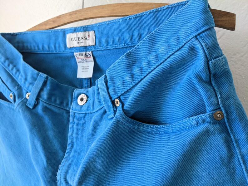 Puede incluir: Un par de pantalones vaqueros azules con una etiqueta de Guess Jeans. Los vaqueros tienen un lavado azul claro y un estilo cl&aacute;sico de cinco bolsillos.