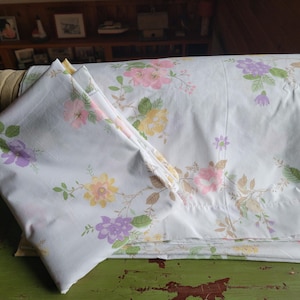 May include: A set of white floral bed sheets with a pink, yellow, and purple floral pattern. The sheets are folded and stacked on a green surface.