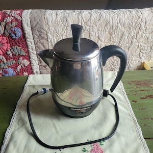 Puede incluir: Una cafetera eléctrica Farberware de acero inoxidable con asa y pomo negros. La cafetera se encuentra sobre un mantel bordado verde claro con un diseño floral y de pájaros. Un cable de alimentación negro se extiende desde la base.