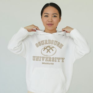 May include: White hoodie with a brown graphic of a sourdough bread loaf and the text "Sourdough University" with the words "Breaducated" underneath.