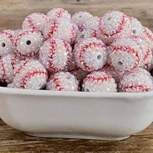 May include: A white ceramic bowl filled with baseball-themed beads. The beads are white with red stitching and covered in iridescent rhinestones. The beads have a hole through the center.