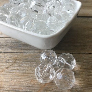 May include: A white bowl filled with clear faceted glass beads. Four additional beads are scattered on a wooden surface.